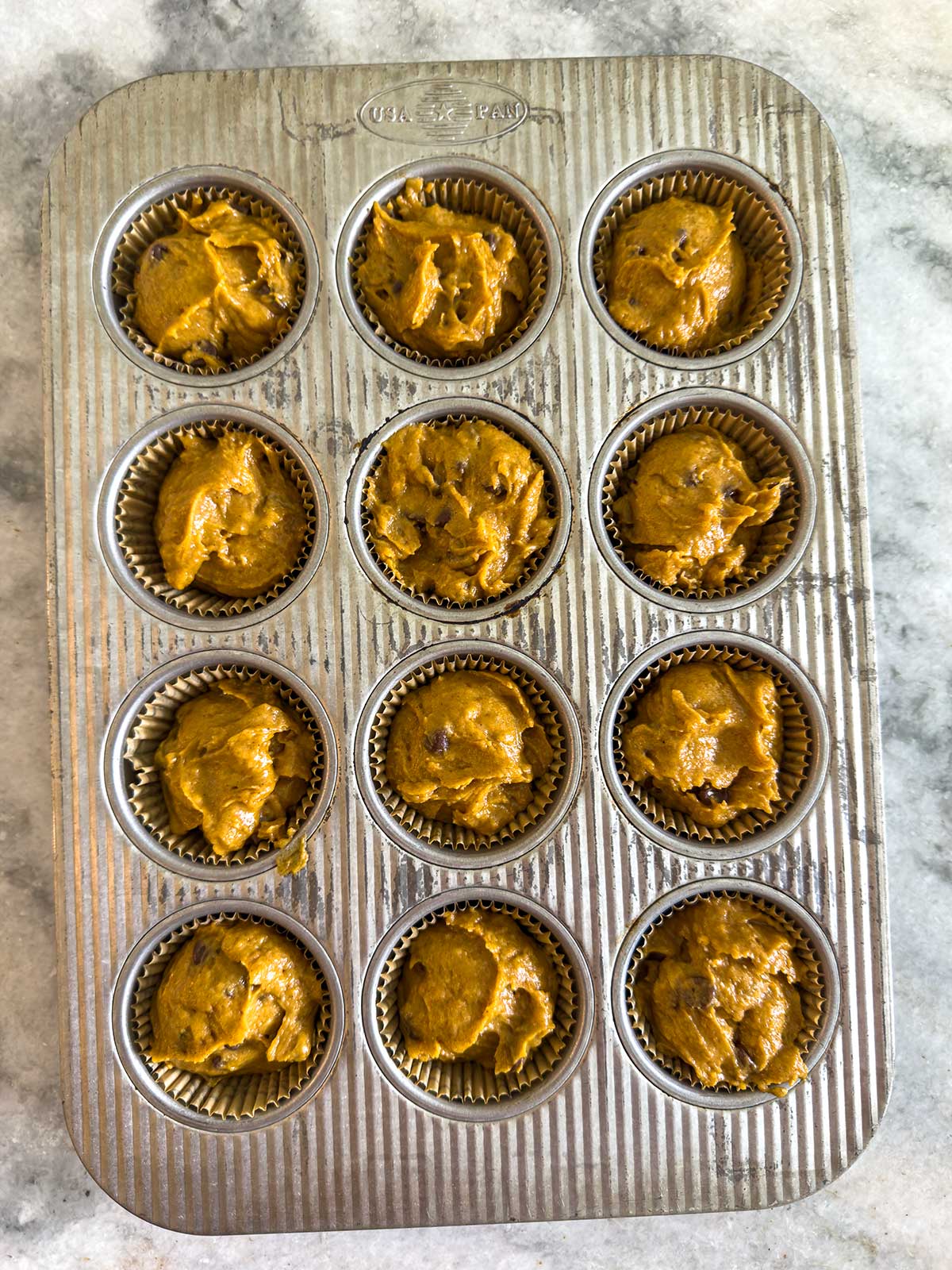 A metal muffin tin with 12 cups, each lined with a paper liner and filled with unbaked, brownish-orange muffin batter, sits on a gray marble surface.