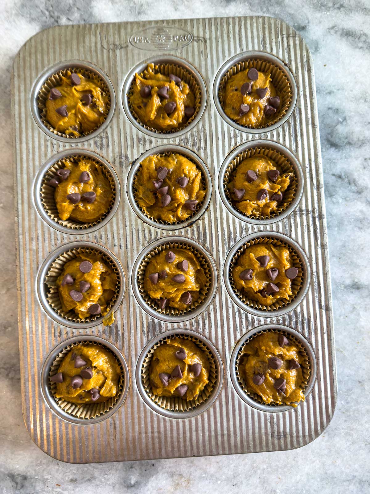 A muffin tin filled with twelve paper liners, each containing unbaked orange batter topped with chocolate chips, sits on a gray marble surface.
