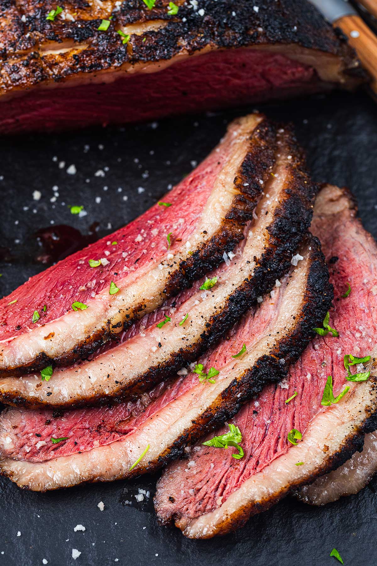 Four slices of medium-rare seasoned picanha steak with a charred crust, sprinkled with coarse salt and chopped herbs, are arranged on a dark slate surface with part of a larger steak piece visible.