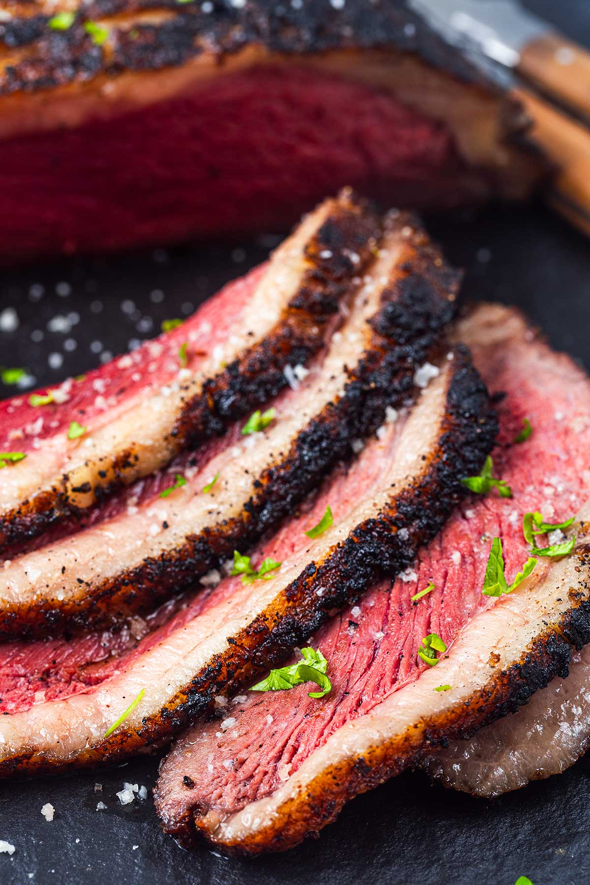 Sliced, medium-rare picanha steak with a charred crust, sprinkled with sea salt and chopped parsley, displayed on a dark surface.