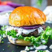 A close-up of a blue cheese burger with a shiny brioche bun, fresh green leaf lettuce, a beef patty, sliced red onion, and creamy white sauce, with some sauce dripping onto the slate surface.
