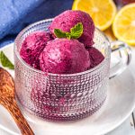 A glass cup filled with scoops of purple berry frozen yogurt, garnished with a mint leaf, sits on a white plate. A wooden spoon and sliced lemons are beside it, with a blue cloth in the background.