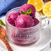 A glass cup filled with scoops of purple berry frozen yogurt, garnished with a mint leaf, sits on a white plate. A wooden spoon and sliced lemons are beside it, with a blue cloth in the background.