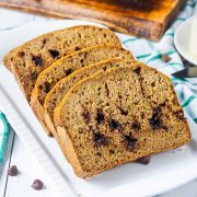 Four slices of zucchini bread with chocolate chips are arranged on a white plate, with a green and white checkered towel and a wooden board in the background.
