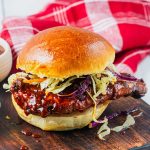A barbecue chicken sandwich with coleslaw on a shiny bun sits on a wooden board, with a red and white cloth in the background.