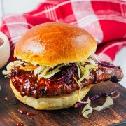 A barbecue chicken sandwich with coleslaw on a shiny bun sits on a wooden board, with a red and white cloth in the background.