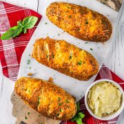Three cheesy garlic breads topped with herbs are on parchment paper over a wooden board. A bowl of parmesan slices and fresh basil leaves are nearby, all placed on a red plaid cloth on a white wooden surface.
