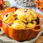 Pumpkin-shaped bowl filled with scoops of orange ice cream, topped with caramel sauce and pecans, placed on a beige cloth with autumn decorations in the background.