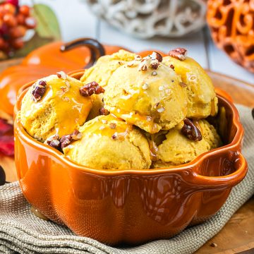 Pumpkin-shaped bowl filled with scoops of orange ice cream, topped with caramel sauce and pecans, placed on a beige cloth with autumn decorations in the background.