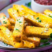 Crispy polenta fries topped with grated cheese and chopped parsley on a plate, with a bowl of dipping sauce in the background.