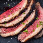 Four slices of medium-rare grilled picanha steak with a seared crust, sprinkled with coarse salt and chopped herbs, arranged on a dark surface.