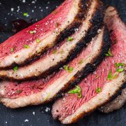 Four slices of medium-rare grilled picanha steak with a seared crust, sprinkled with coarse salt and chopped herbs, arranged on a dark surface.