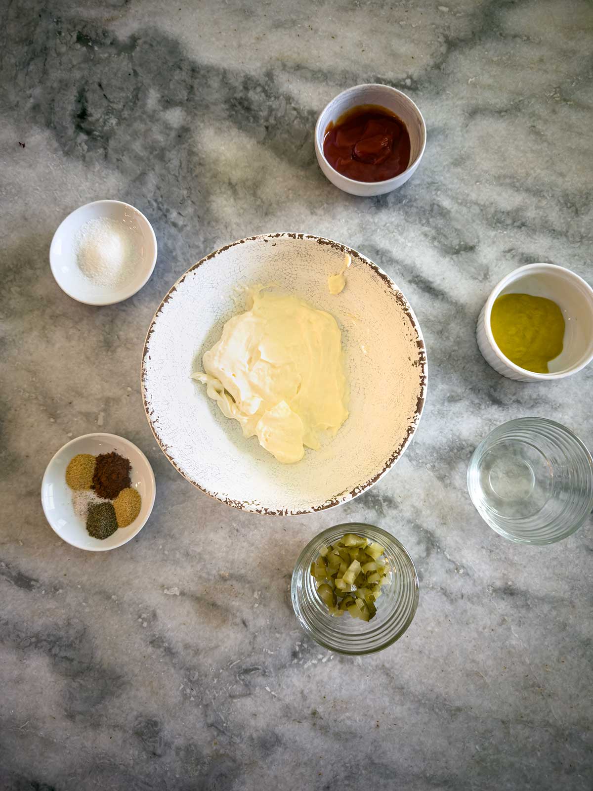 A bowl of mayonnaise is surrounded by small bowls containing ketchup, mustard, pickles, spices, sugar, and water&mdash;all the classic ingredients for crafting a delicious homemade smashburger sauce&mdash;neatly arranged on a gray marble surface.