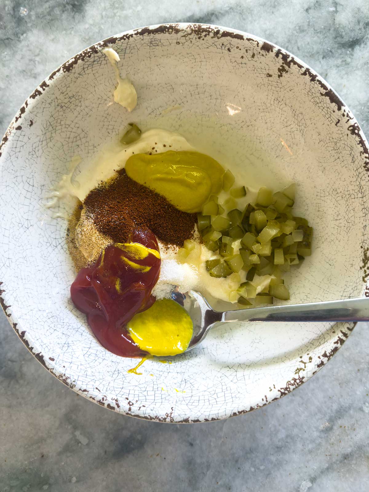 A bowl of smashburger sauce, made with ketchup, mustard, chopped pickles, mayonnaise, and spices, with a spoon resting inside—all on a gray marble surface.