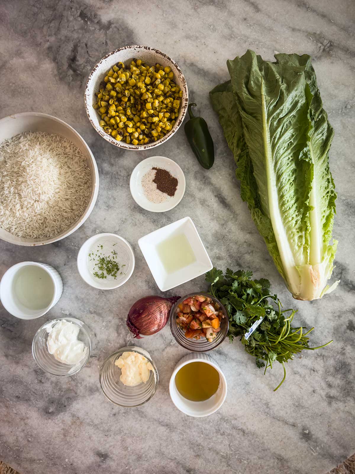 A marble countertop displays ingredients for a street corn chicken rice bowl in bowls and jars: rice, corn, jalapeño, lettuce, cilantro, pico de gallo, red onion, olive oil, white sauce, mayonnaise, salt, pepper, lime zest, and vinegar.