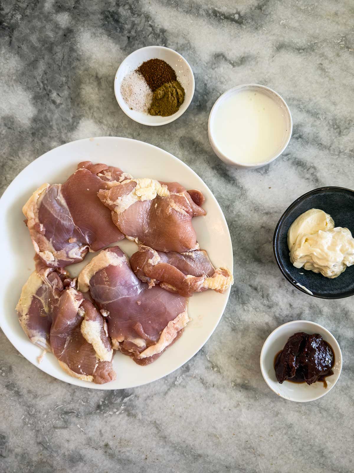 Raw chicken thighs on a white plate are surrounded by spices, buttermilk, mayonnaise, and a dark paste—perfect for creating a flavorful street corn chicken rice bowl on a gray marble surface.