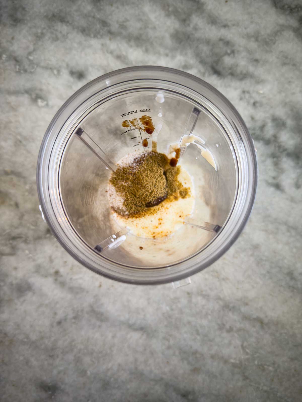 Overhead view of a blender cup containing white liquid and piles of brown and greenish spices for a street corn chicken rice bowl, all sitting on a gray marble countertop.