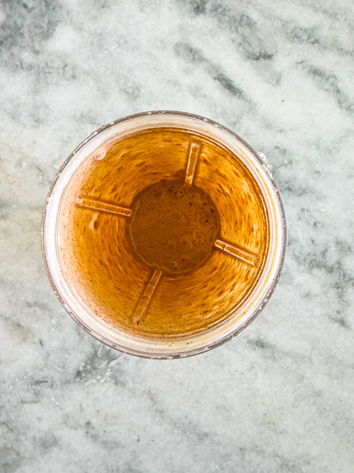 A top-down view of a glass filled with a brownish-golden liquid, possibly tea or coffee, sits on a light gray marble surface beside a street corn chicken rice bowl. The glass has visible blender blade marks on the sides.