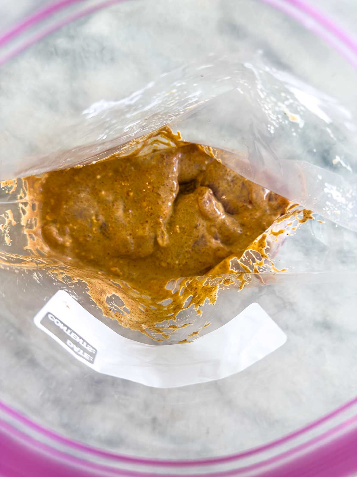 A close-up view of a resealable plastic bag containing a thick, orange-brown marinade coating pieces of raw meat, perfect for a flavorful street corn chicken rice bowl, placed on a light-colored surface.