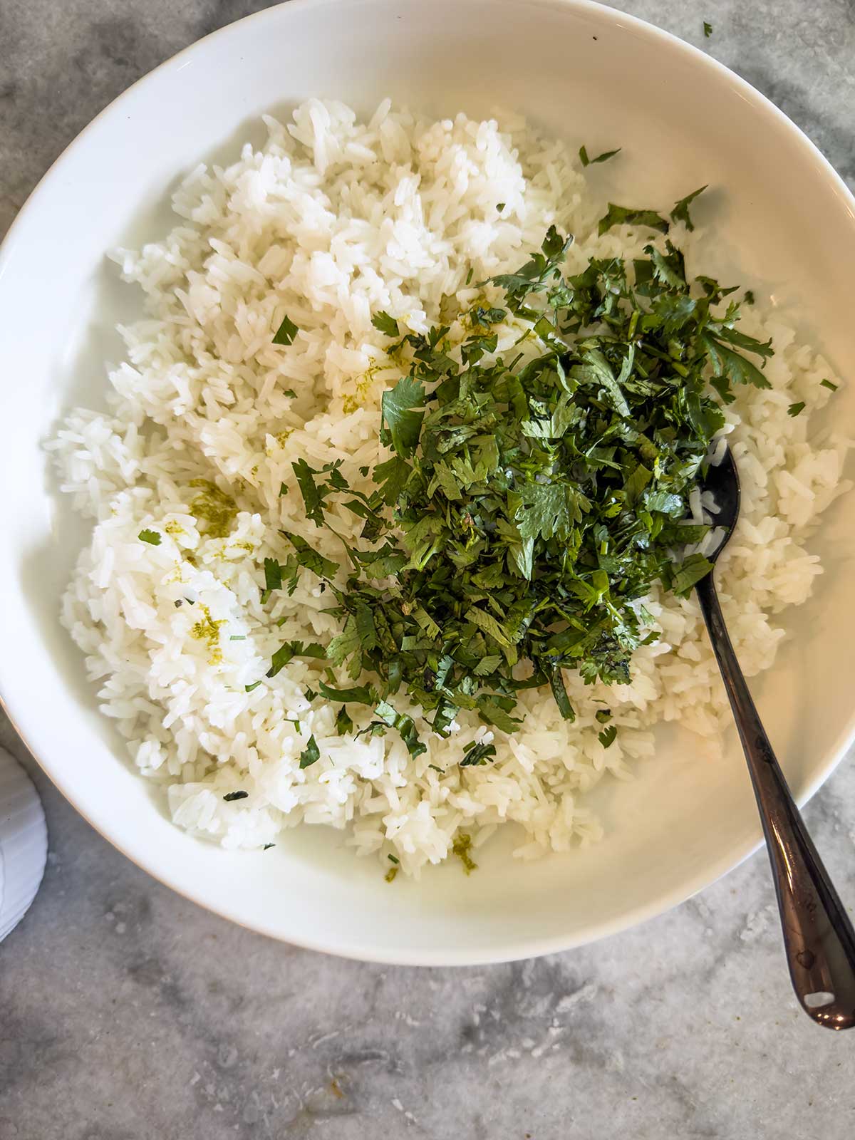 A white bowl filled with cooked white rice, topped with chopped fresh herbs—likely cilantro—and reminiscent of a street corn chicken rice bowl, with a spoon resting inside on a gray countertop.