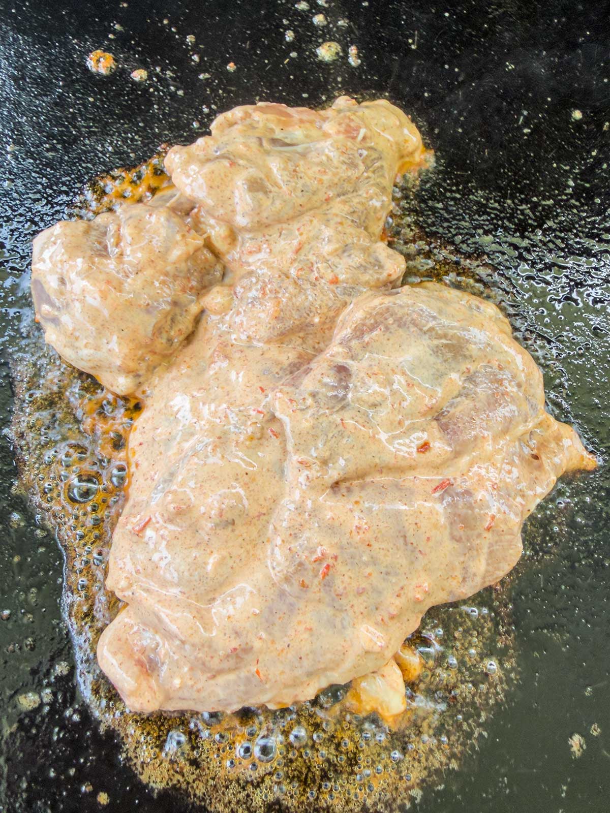 A piece of marinated meat sizzles on a hot, oily pan, producing bubbles around the edges as it cooks—perfect for topping a vibrant street corn chicken rice bowl.