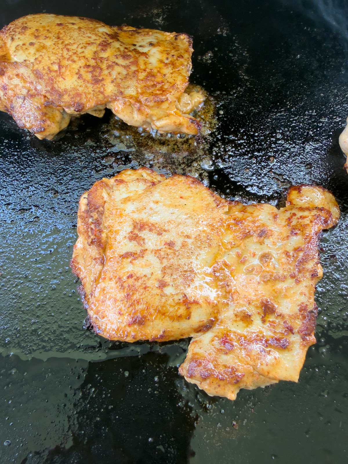 Two pieces of seasoned and cooked chicken breast sizzle on a greased, dark stovetop—golden brown with crisp edges—perfect for topping a street corn chicken rice bowl.