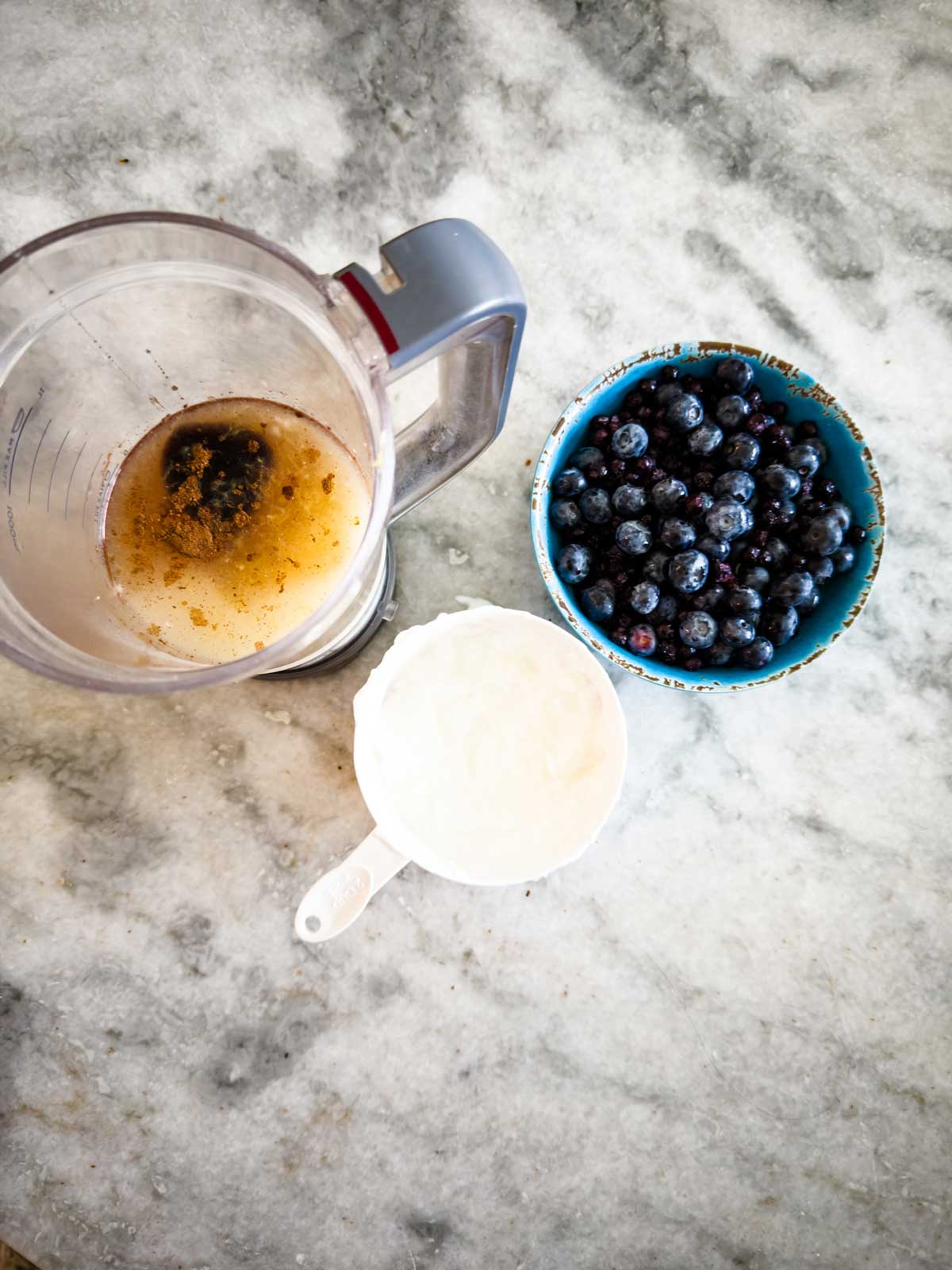 A blender jar with brown liquid and spices, a blue bowl of blueberries, and a white measuring cup filled with yogurt sit on a marble countertop, ready to be transformed into creamy blueberry frozen yogurt.