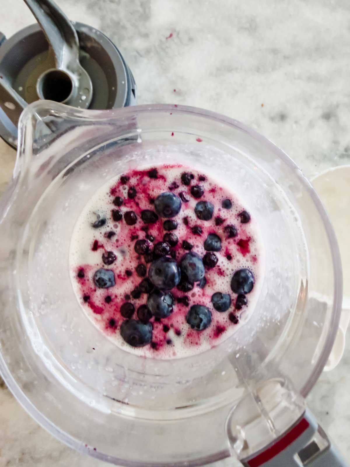 A blender container with blueberries and white liquid, possibly milk or yogurt, inside—suggesting the start of a homemade blueberry frozen yogurt—viewed from above on a marble countertop. Blender parts are visible nearby.