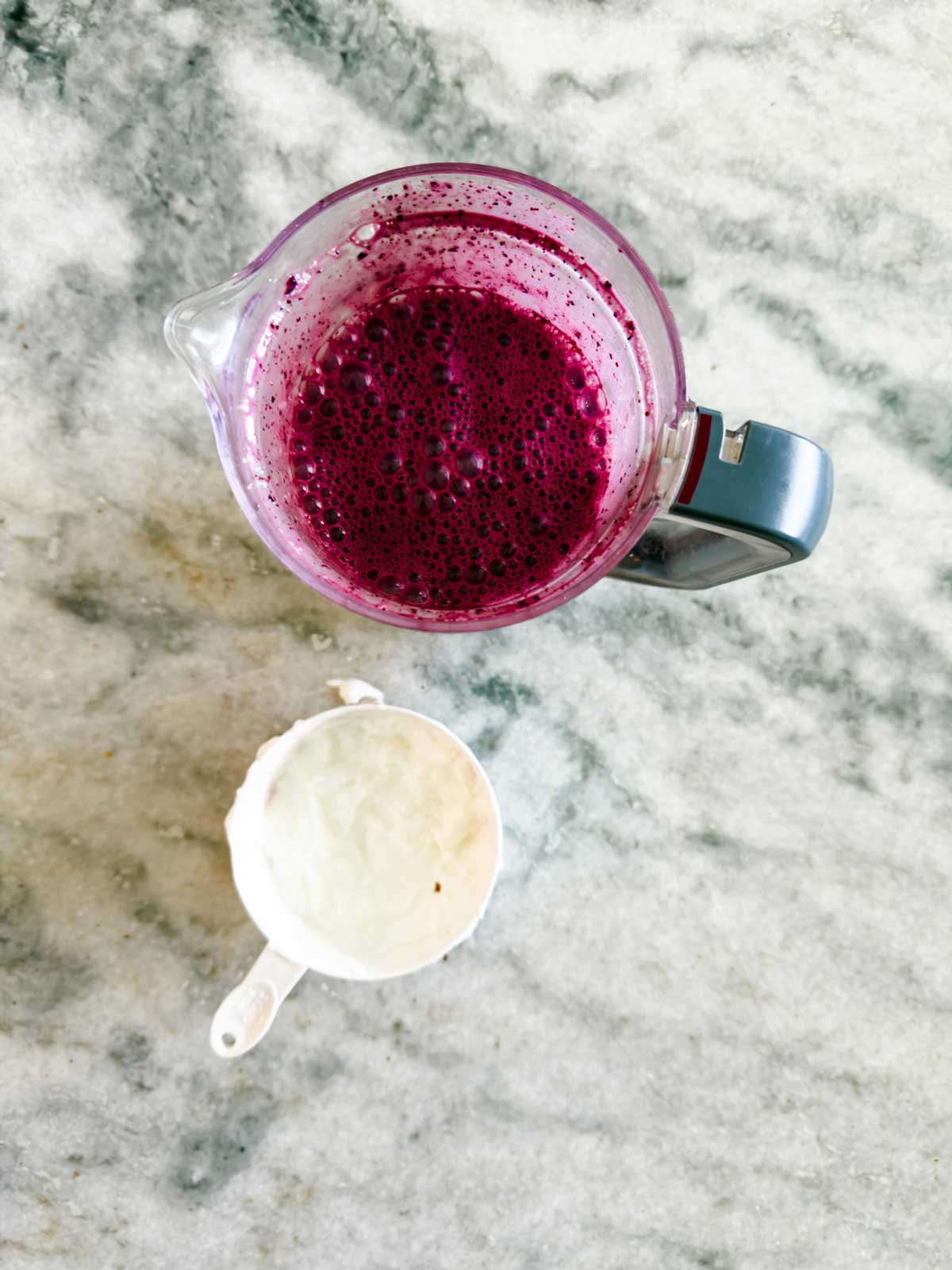 A blender with vibrant purple blueberry frozen yogurt smoothie and a white measuring cup with a small amount of white ingredient sit on a marble countertop.