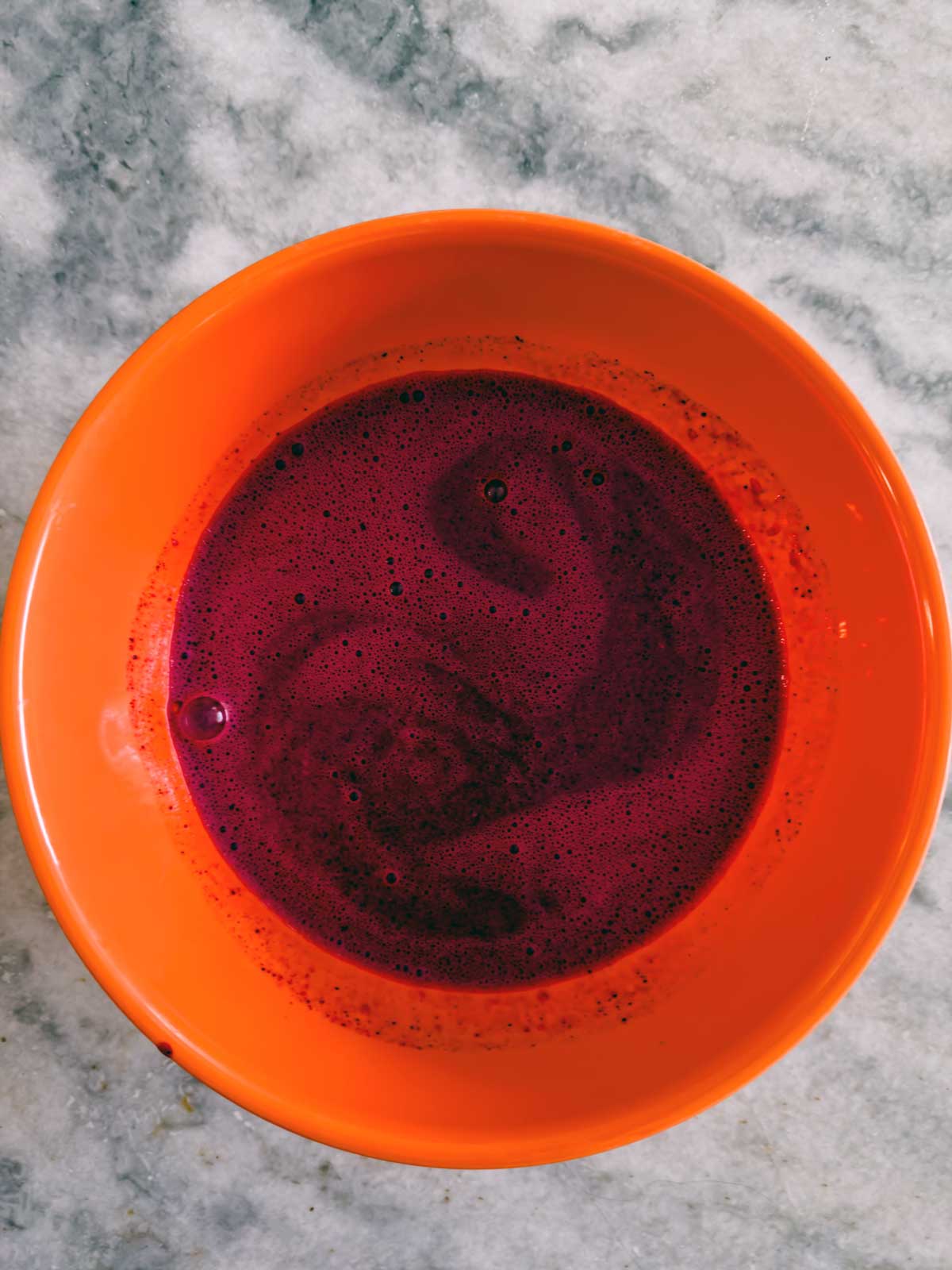 An orange bowl filled with dark purple liquid, possibly blueberry frozen yogurt or soup, sits on a gray marble surface.