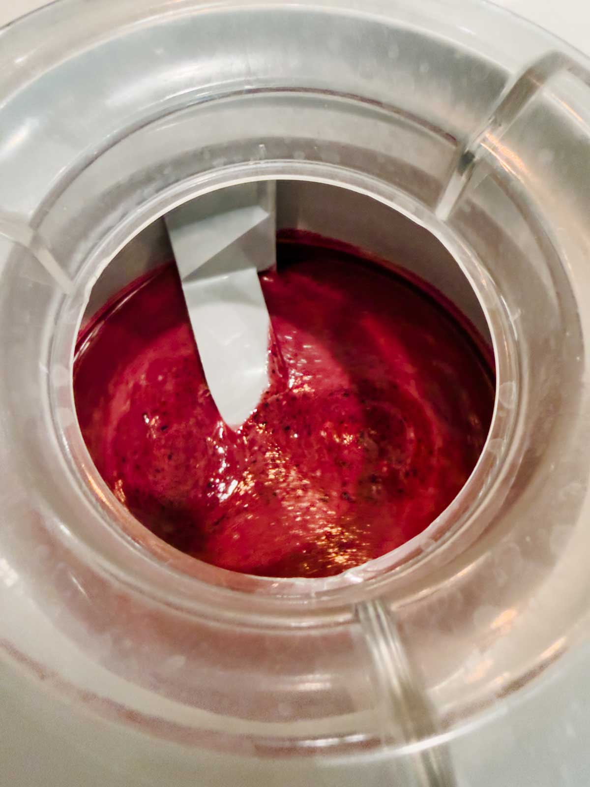 A close-up view inside an ice cream maker churning a dark red mixture, likely a fruit sorbet, gelato, or blueberry frozen yogurt, with the paddle visible in the center.
