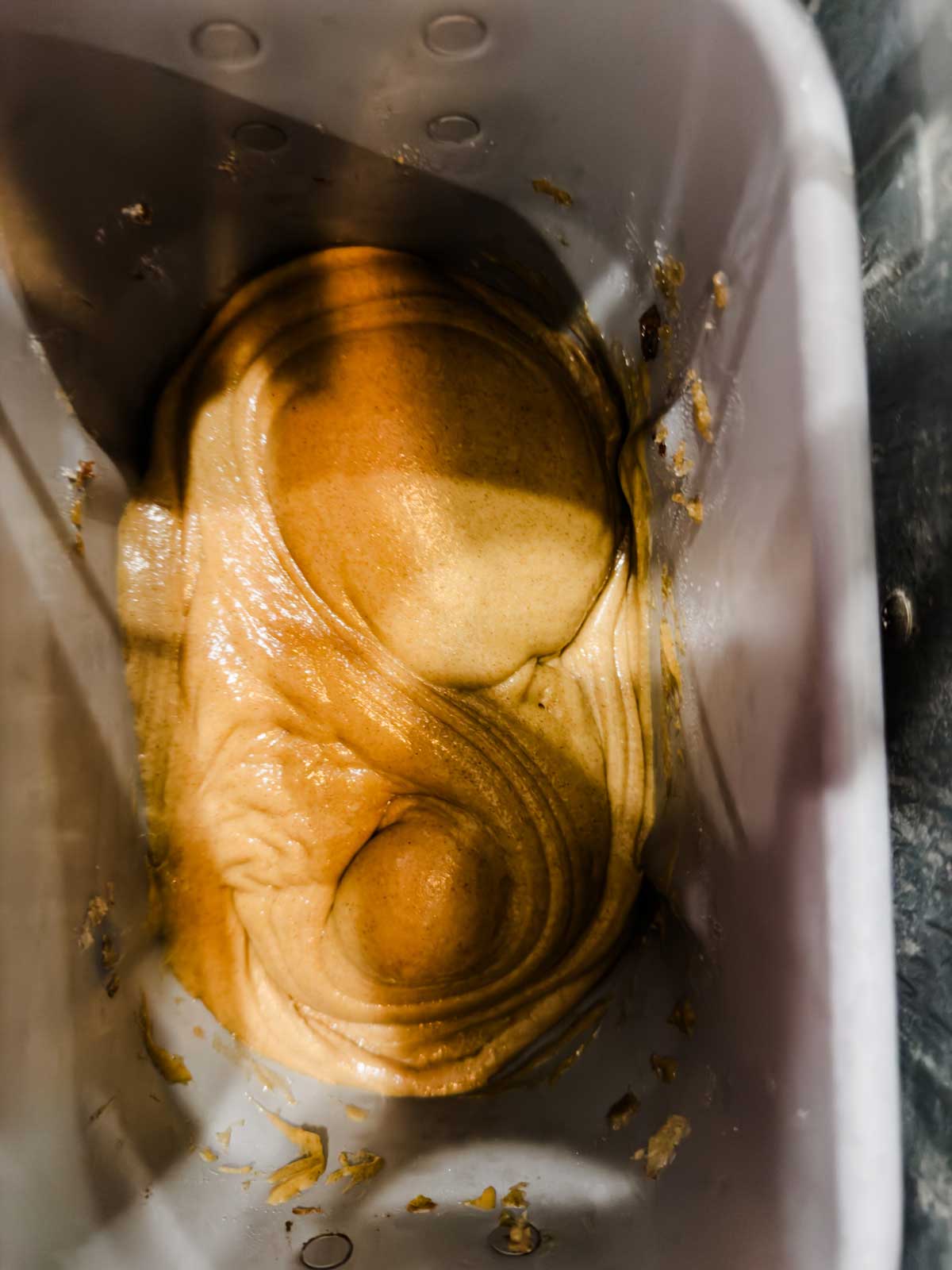 Sticky, golden brown dough being kneaded inside a bread machine pan, viewed from above. The dough has a smooth, glossy texture and is swirling into a soft mound against the sides of the container.