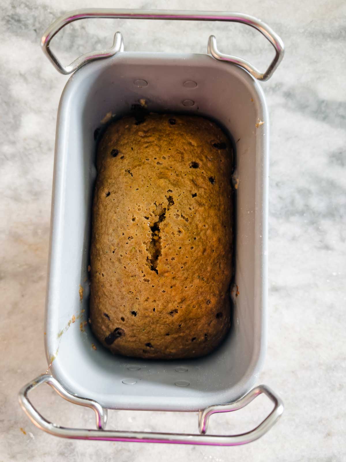 A loaf of baked banana bread with chocolate chips sits in a metal loaf pan on a light marble countertop.