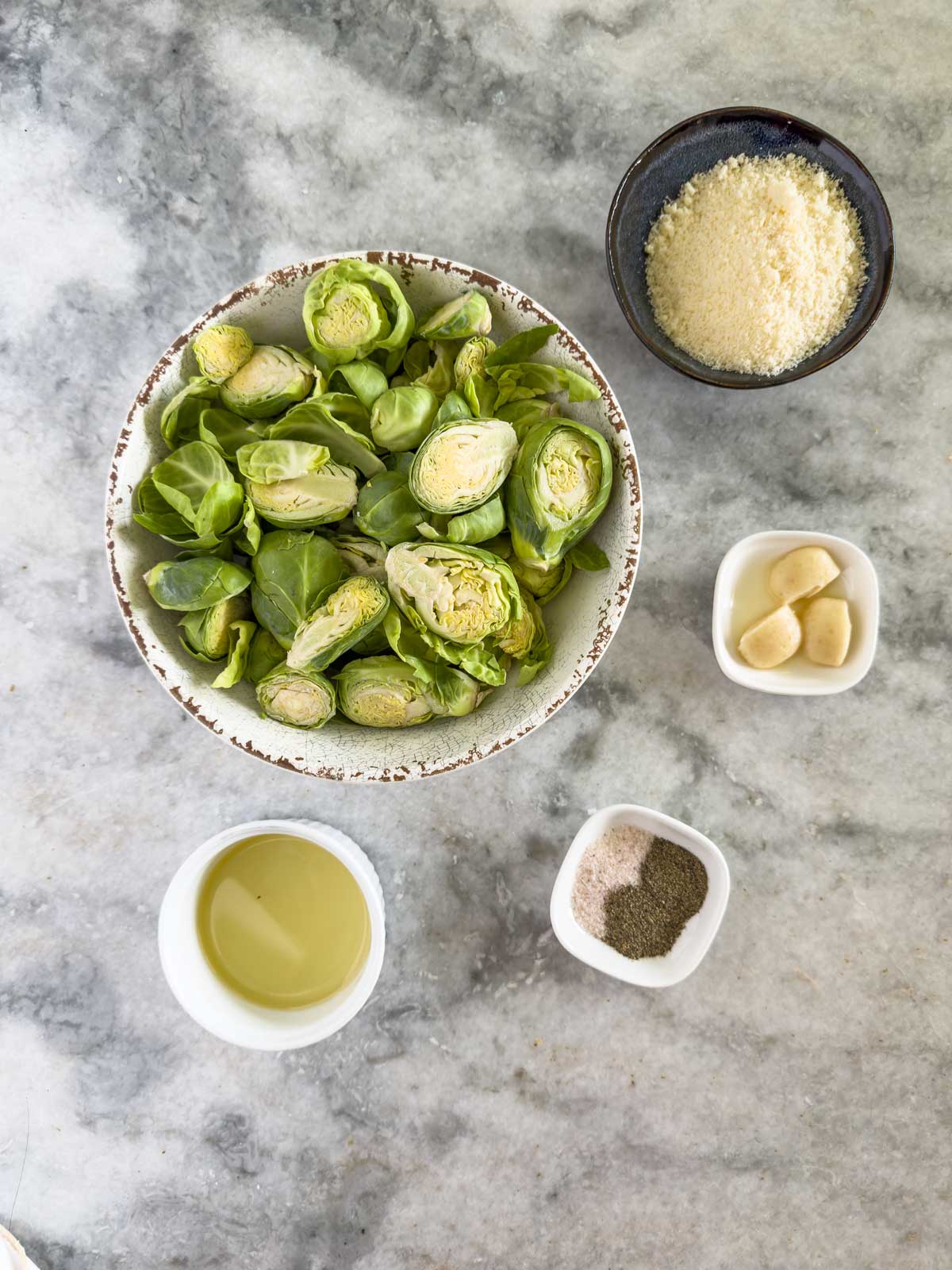 A bowl of halved Brussels sprouts, a small bowl of grated cheese for a rich parmesan flavor, garlic cloves, a cup of oil, and a dish of salt and pepper are arranged on a gray marble surface—everything you need to create flavorful garlic parmesan Brussels sprouts.