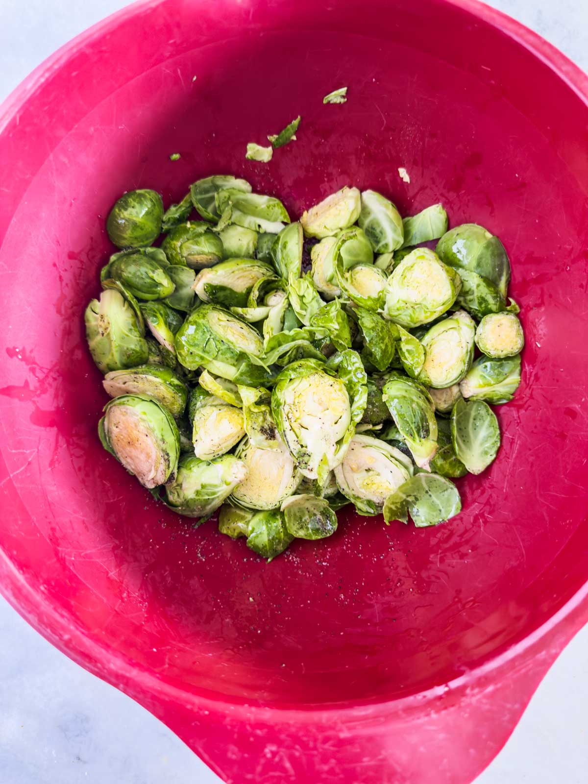 Sliced Brussels sprouts seasoned with salt and pepper in a large red mixing bowl, ready to be cooked into delicious garlic parmesan brussels sprouts.