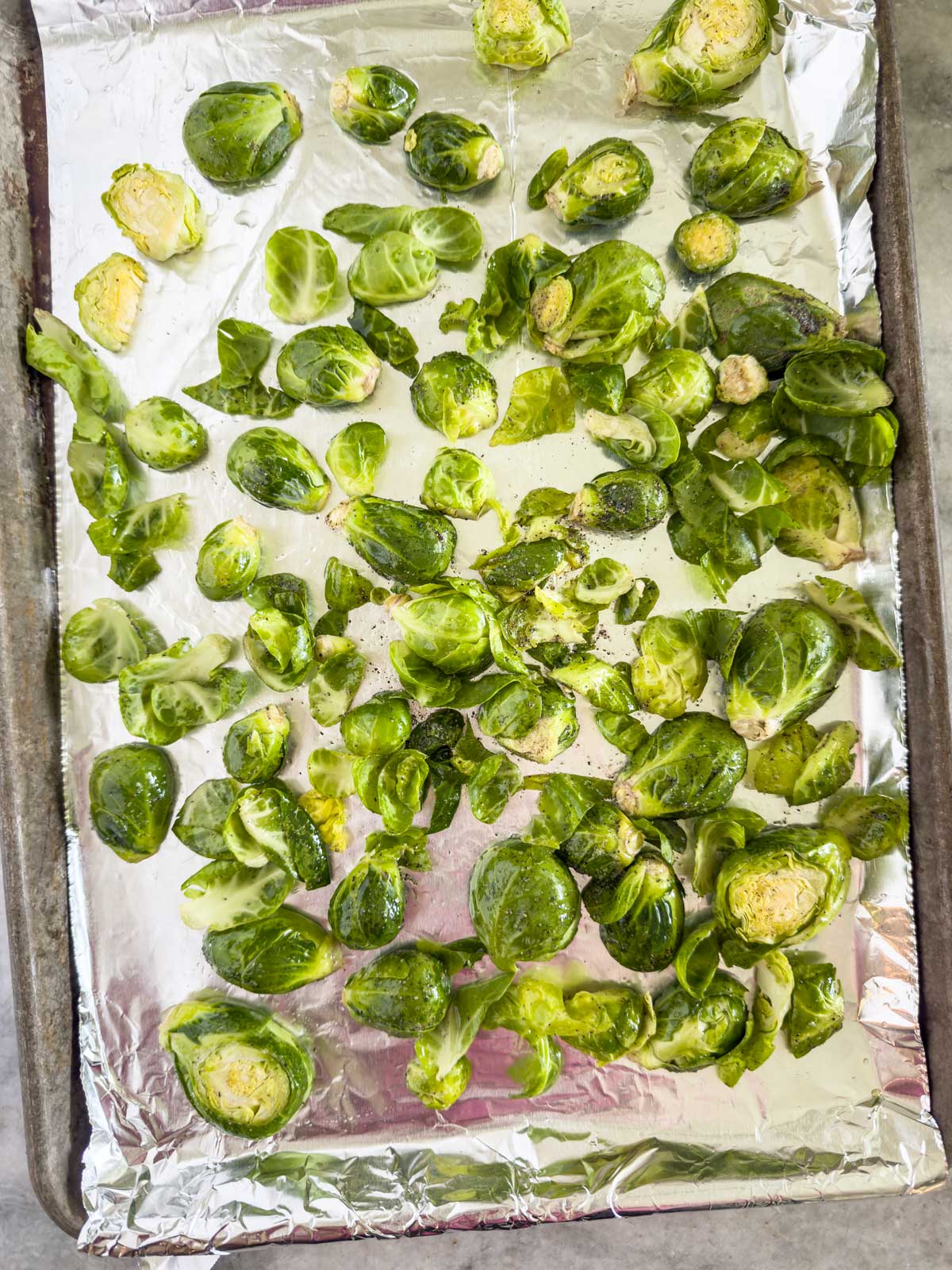 Brussels sprouts, halved and tossed with oil and pepper, are spread out on a foil-lined baking sheet, ready to be roasted in the oven for delicious garlic parmesan Brussels sprouts.