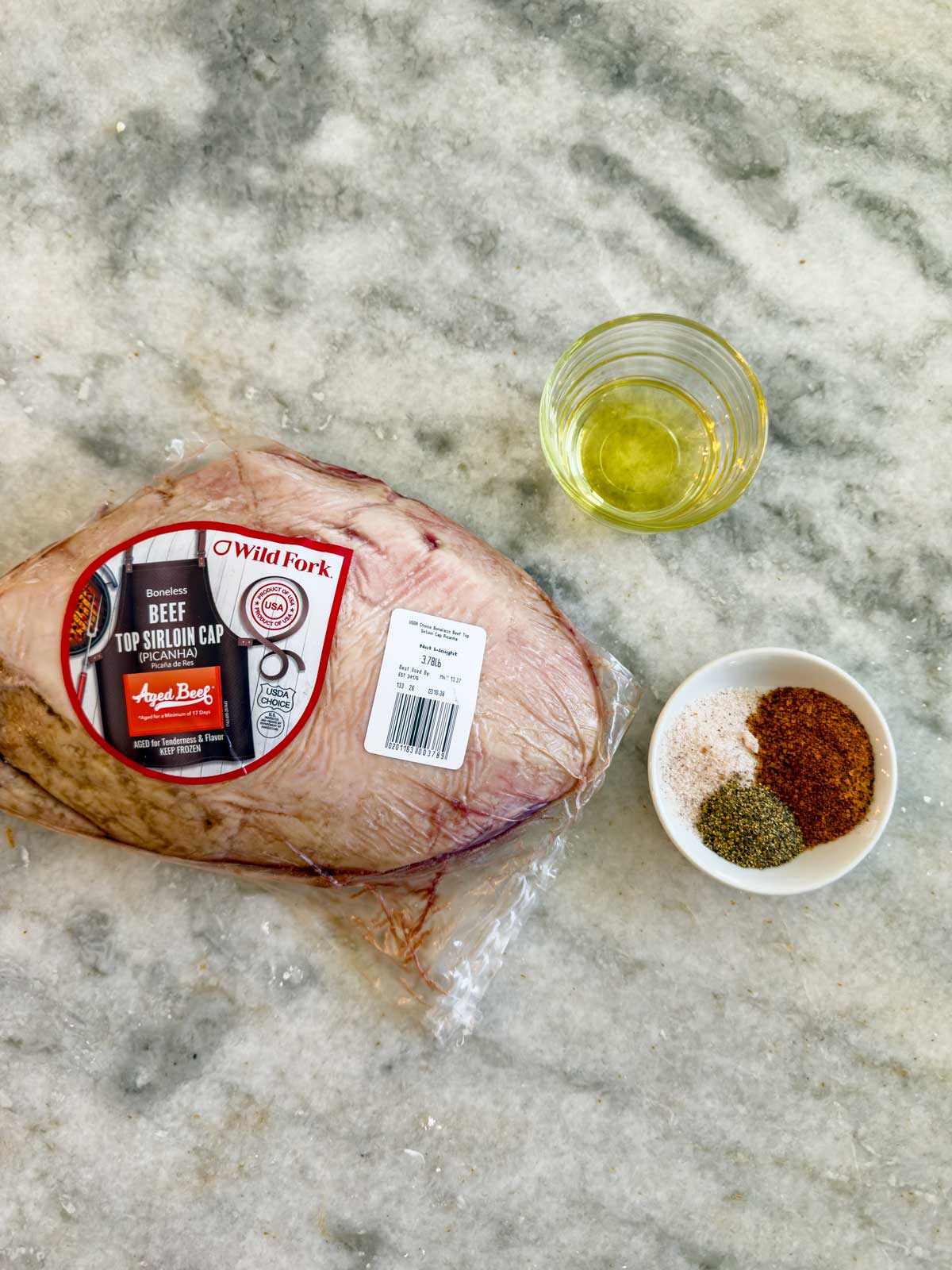 A picanha, vacuum-sealed for freshness, sits on a marble surface next to a glass of oil and a small white bowl containing a mix of dry seasonings.