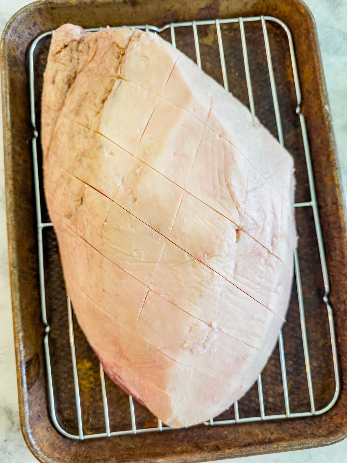 A large raw picanha with a diamond-patterned scored fat cap sits on a wire rack inside a baking tray, reminiscent of the preparation style used for reverse seared picanha.