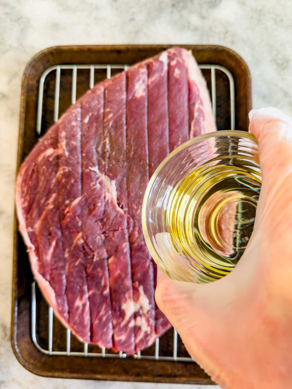 A gloved hand holds a glass cup of oil above a raw, scored reverse seared picanha steak resting on a wire rack in a baking tray.
