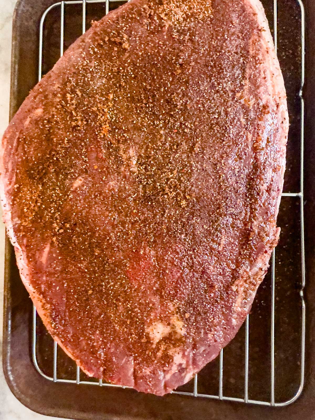 A large, seasoned brisket sits on a wire rack over a baking tray, ready to be cooked in the style of a reverse seared picanha.