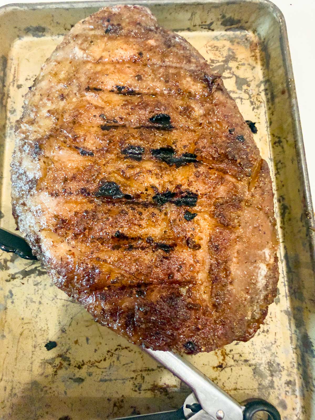 A large, cooked brisket with a browned, seasoned crust rests in a metal baking tray. Grill marks and slight charring are visible on the surface, reminiscent of the techniques used for reverse seared picanha.