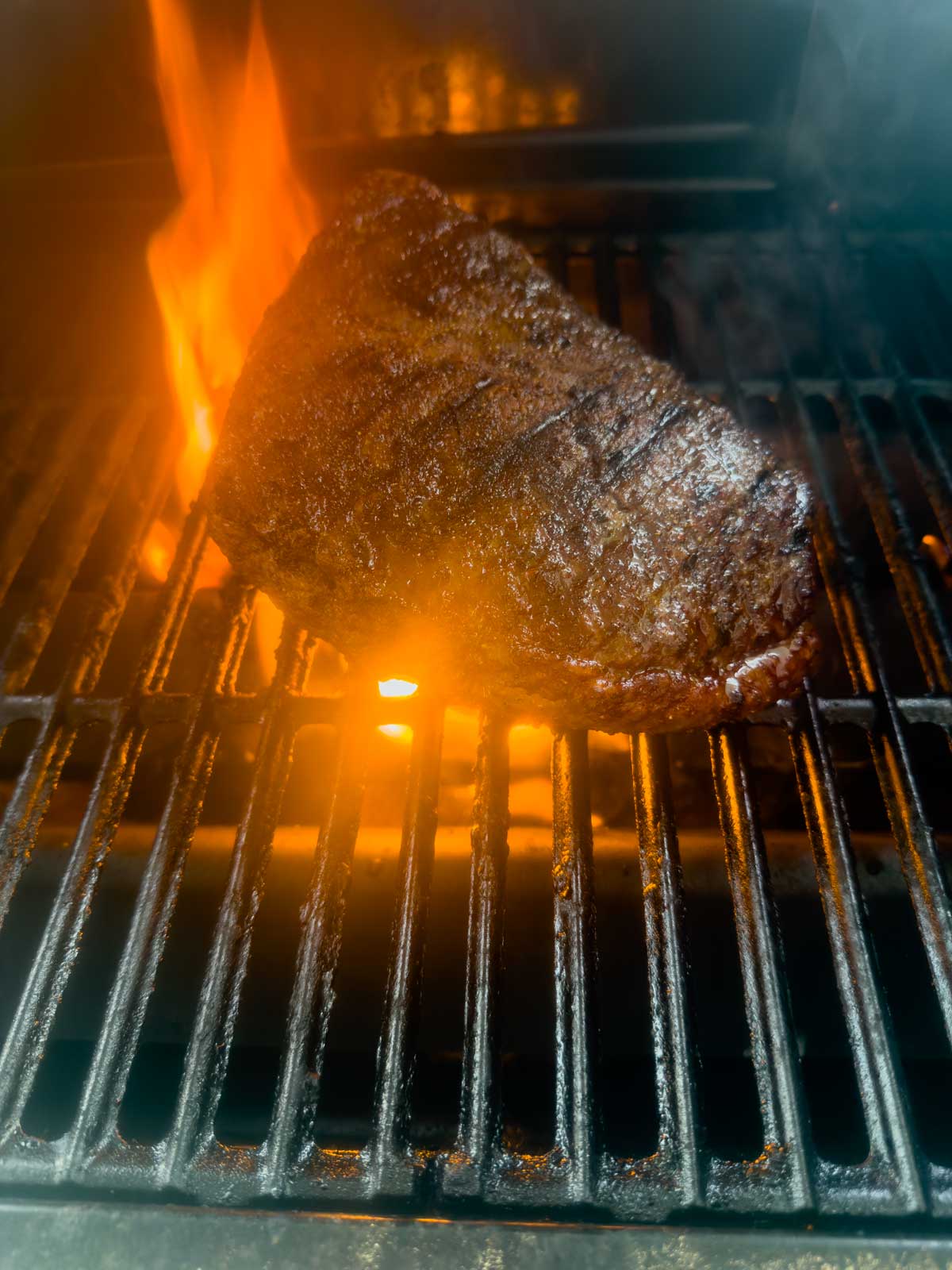 A large piece of seasoned reverse seared picanha is grilling over an open flame on a barbecue, with visible grill marks and fire rising behind it.