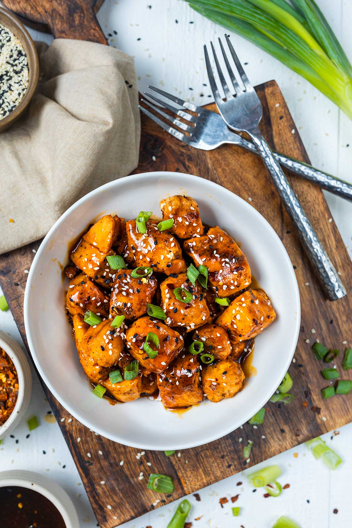 A white bowl filled with glazed salmon cubes topped with sesame seeds and chopped green onions sits on a wooden board, surrounded by forks, sauce, and seasonings.