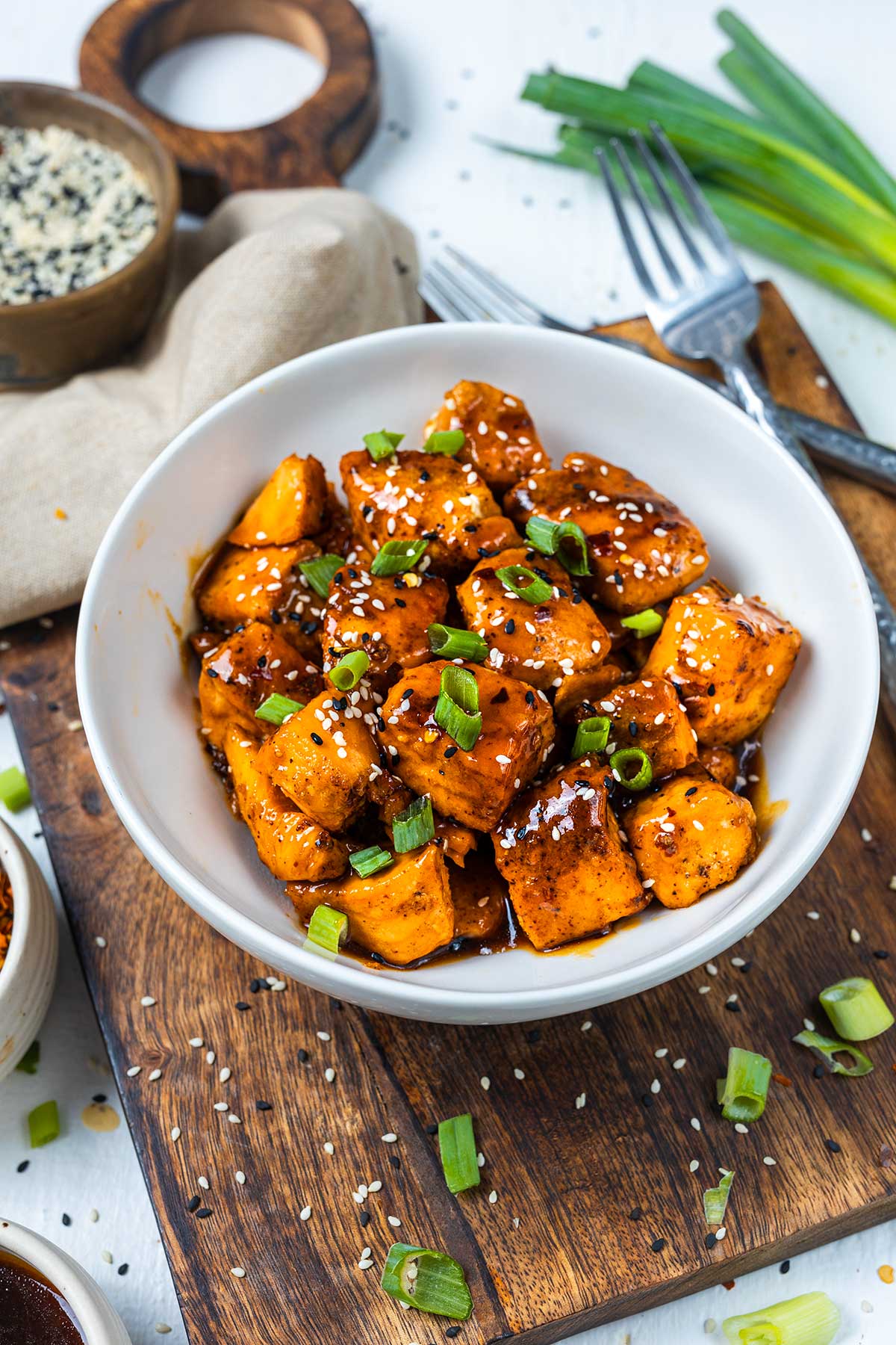 A white bowl filled with saucy salmon cubes garnished with chopped green onions and sesame seeds, placed on a wooden board with utensils and ingredients nearby.