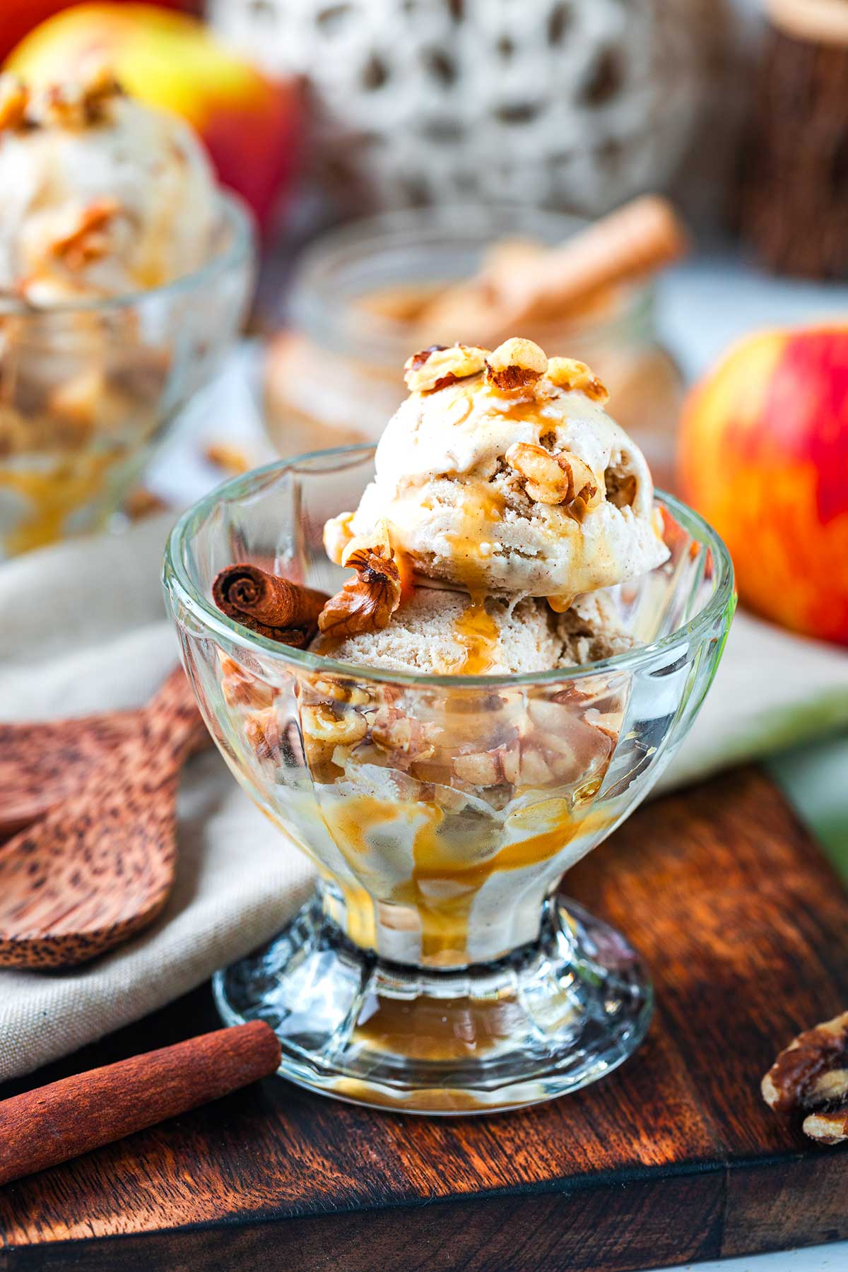 A glass dish with two scoops of ice cream topped with caramel sauce, chopped nuts, and a cinnamon stick. An apple and another dish are in the background on a wooden surface.