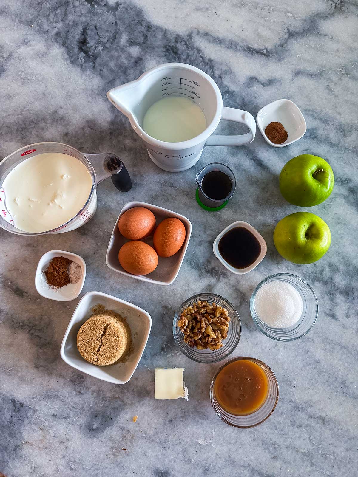 Ingredients for ice cream are arranged on a marble countertop, including eggs, apples, spices, brown and white sugar, milk, pecans, butter, vanilla, caramel sauce, and cream.