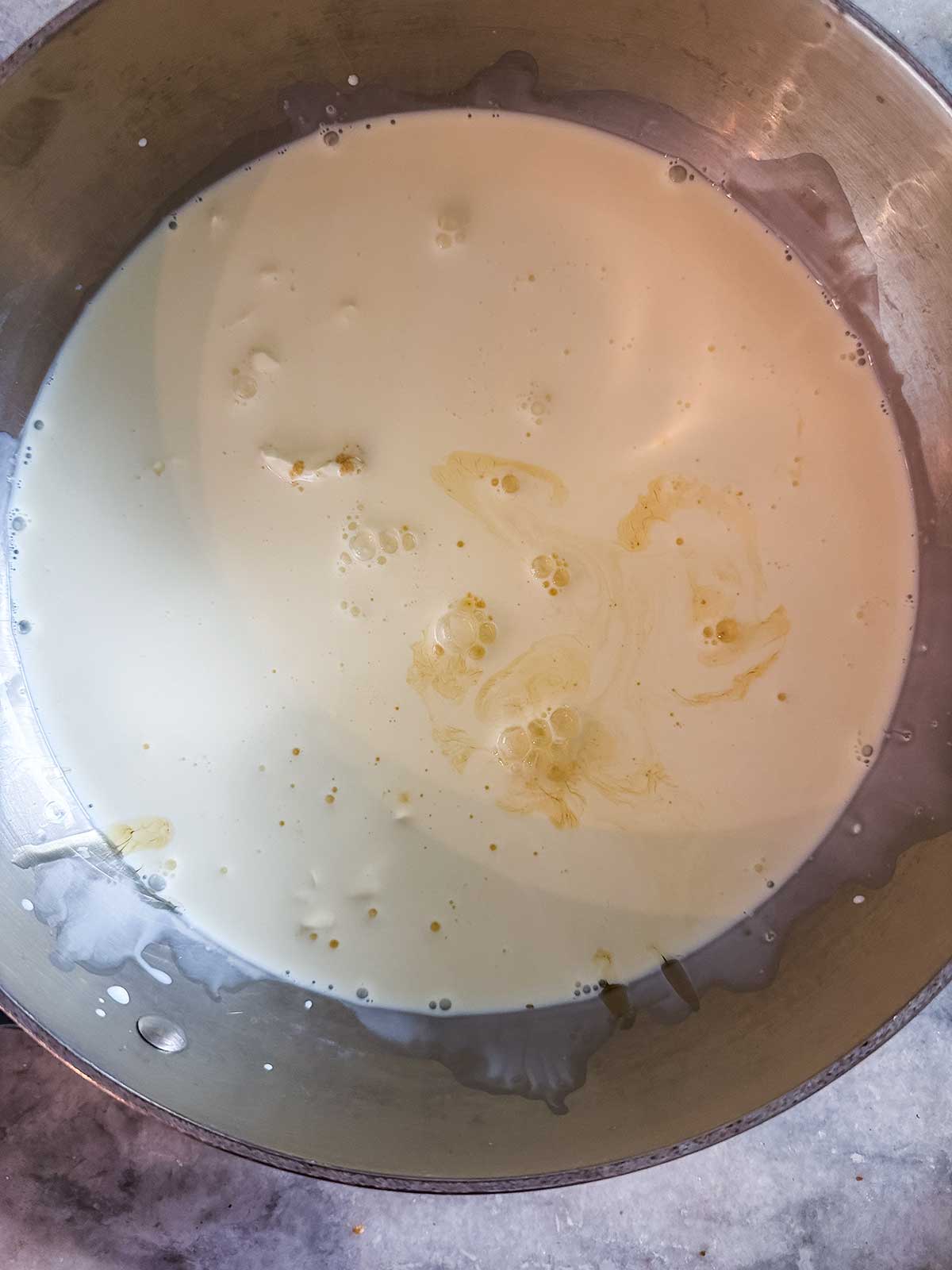 A metal bowl filled with heavy cream, with a few yellowish spots of vanilla extract floating on the surface, sits on a gray countertop.