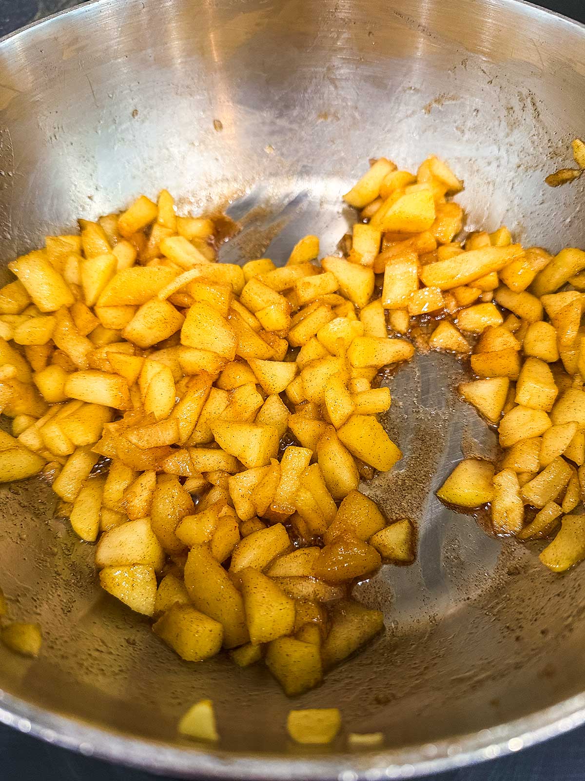 Diced apples cooked with cinnamon and sugar in a stainless steel pot, showing a warm, golden-brown color and a slightly glossy texture.