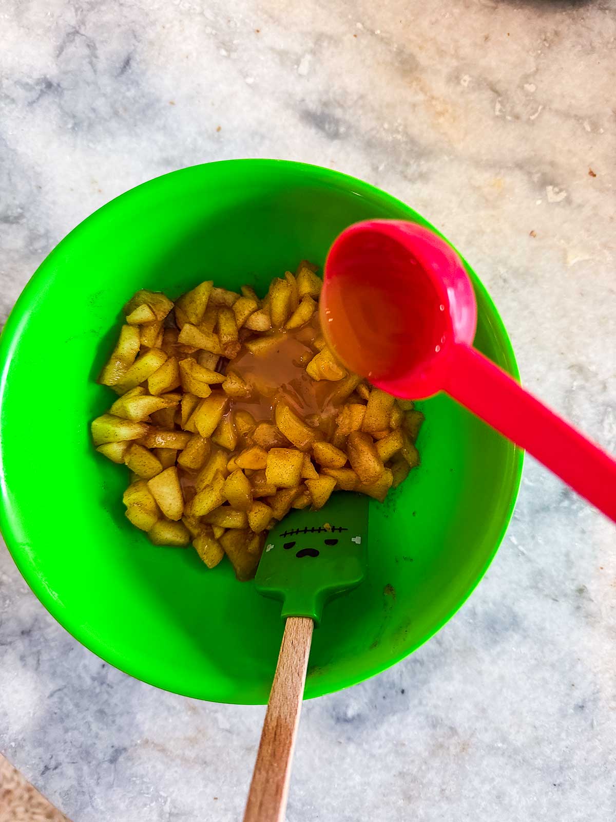 Chopped apples in a green bowl with a spatula. A red measuring spoon is pouring liquid over the apples on a marble countertop.