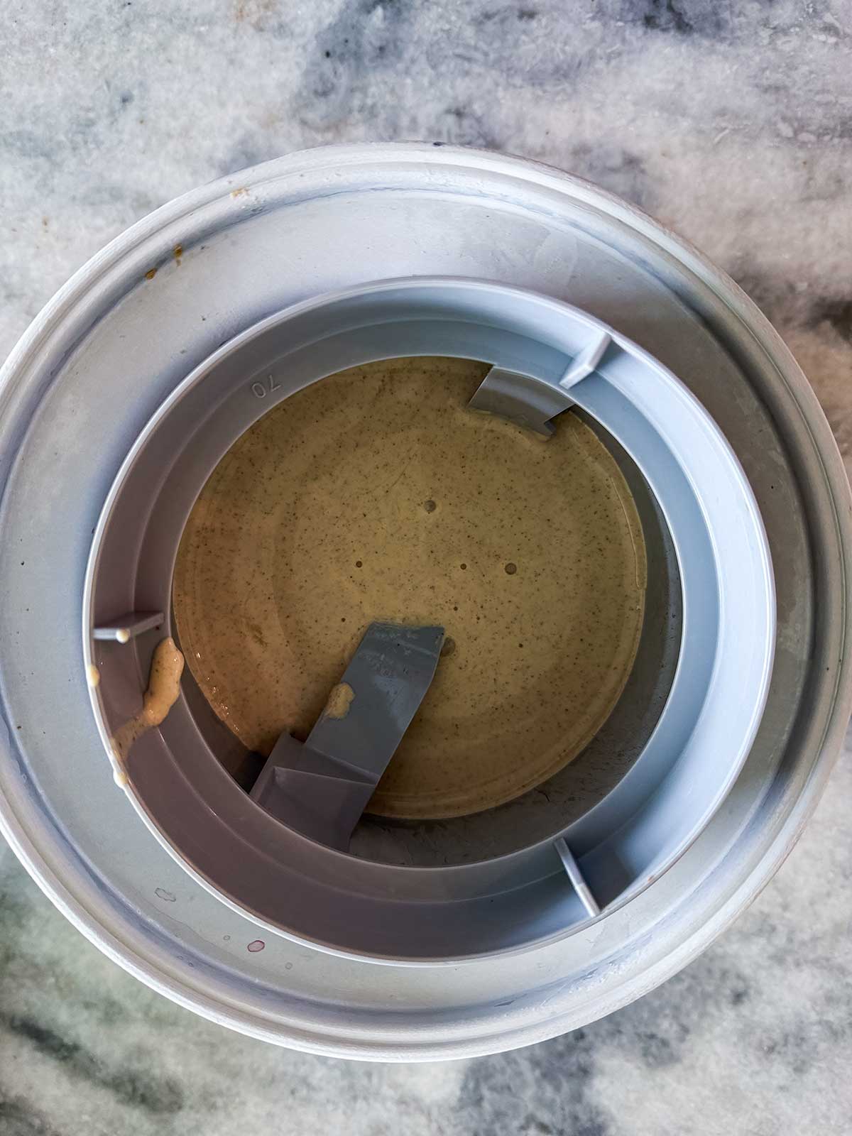Top view of an ice cream maker bowl with light brown ice cream mixture inside, partially churned. The bowl and plastic paddle are visible on a marble countertop.